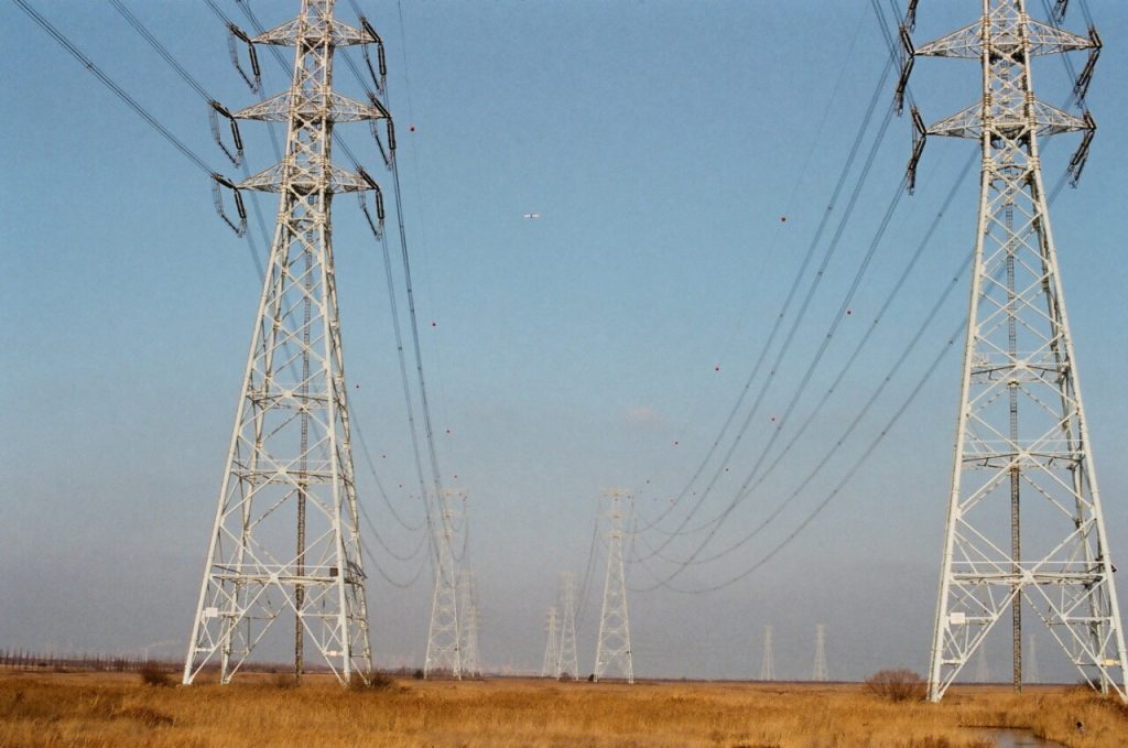 Tall power lines stretch across a dry grassy field under a clear blue sky, resembling a scene from a Regulatory Rollback Rundown.