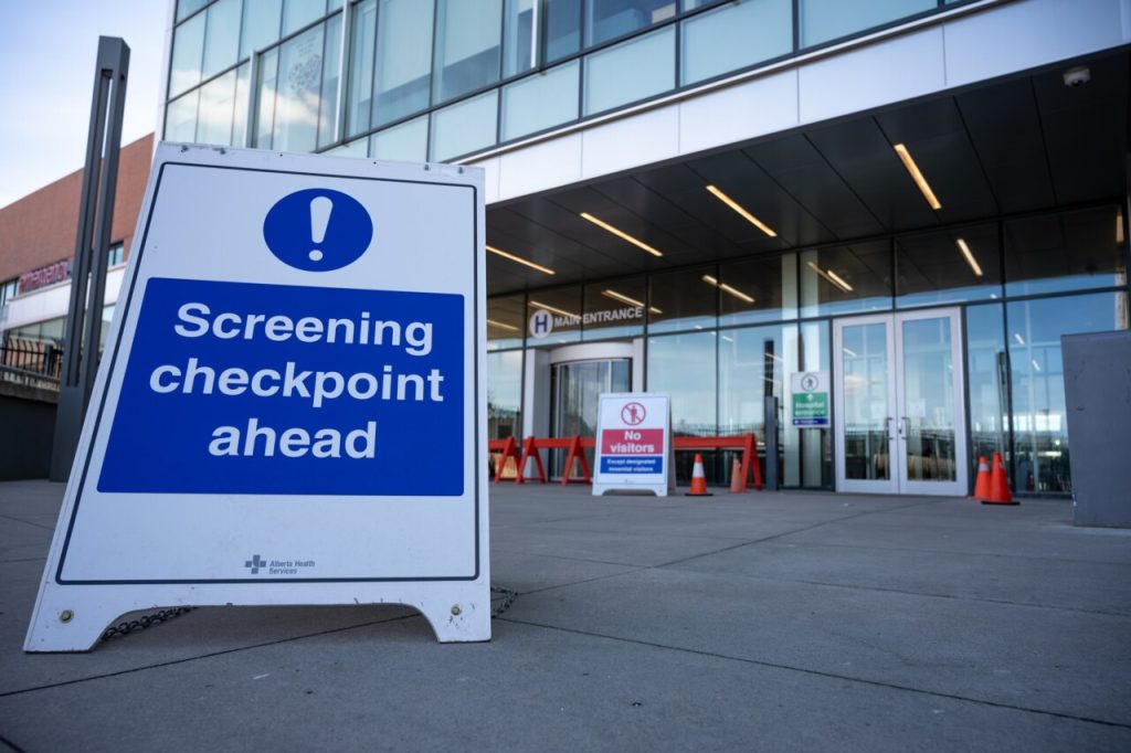 A sign reads Screening checkpoint ahead outside a hospital entrance with glass doors and cones, emphasizing COVID-19 compliance.