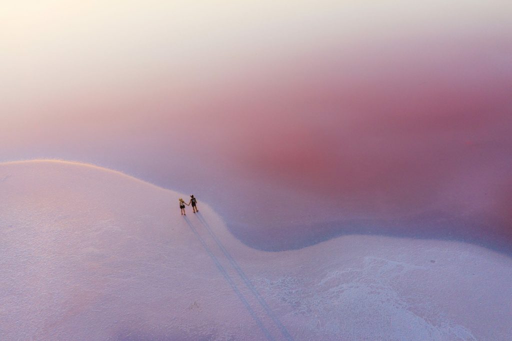Two people walk on a pink and white salt flat, casting long shadows on the textured ground—a striking scene that echoes the harmony between nature and ESG principles promoting EHS and sustainability.