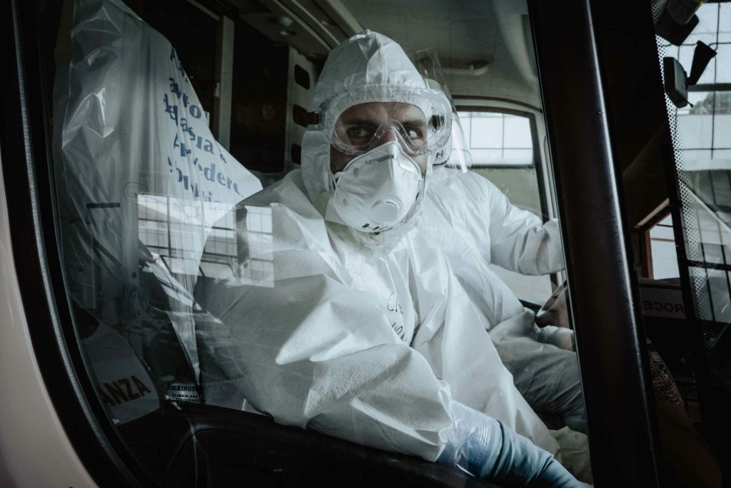 Person in full protective suit and mask sits inside a vehicle, illustrating industrial hygiene practices, while another person sits beside them, both looking out the window.