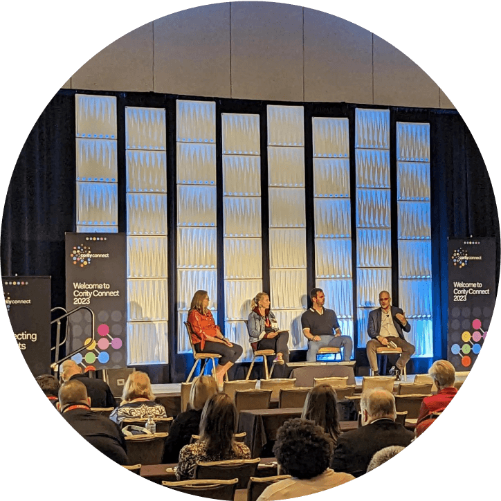 Four people sit on stage in front of an audience, participating in a panel discussion on Sustainability at Cority at a conference. The background features blue-lit panels and banners that read Welcome to Conity Connect 2023.