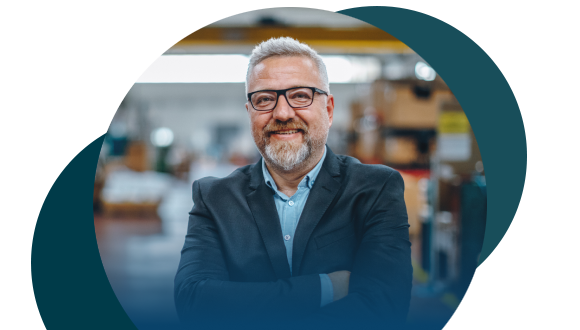 A smiling man with gray hair, beard, and glasses stands with arms crossed in a business suit, in an industrial or warehouse setting with blurred shelves and equipment in the background.