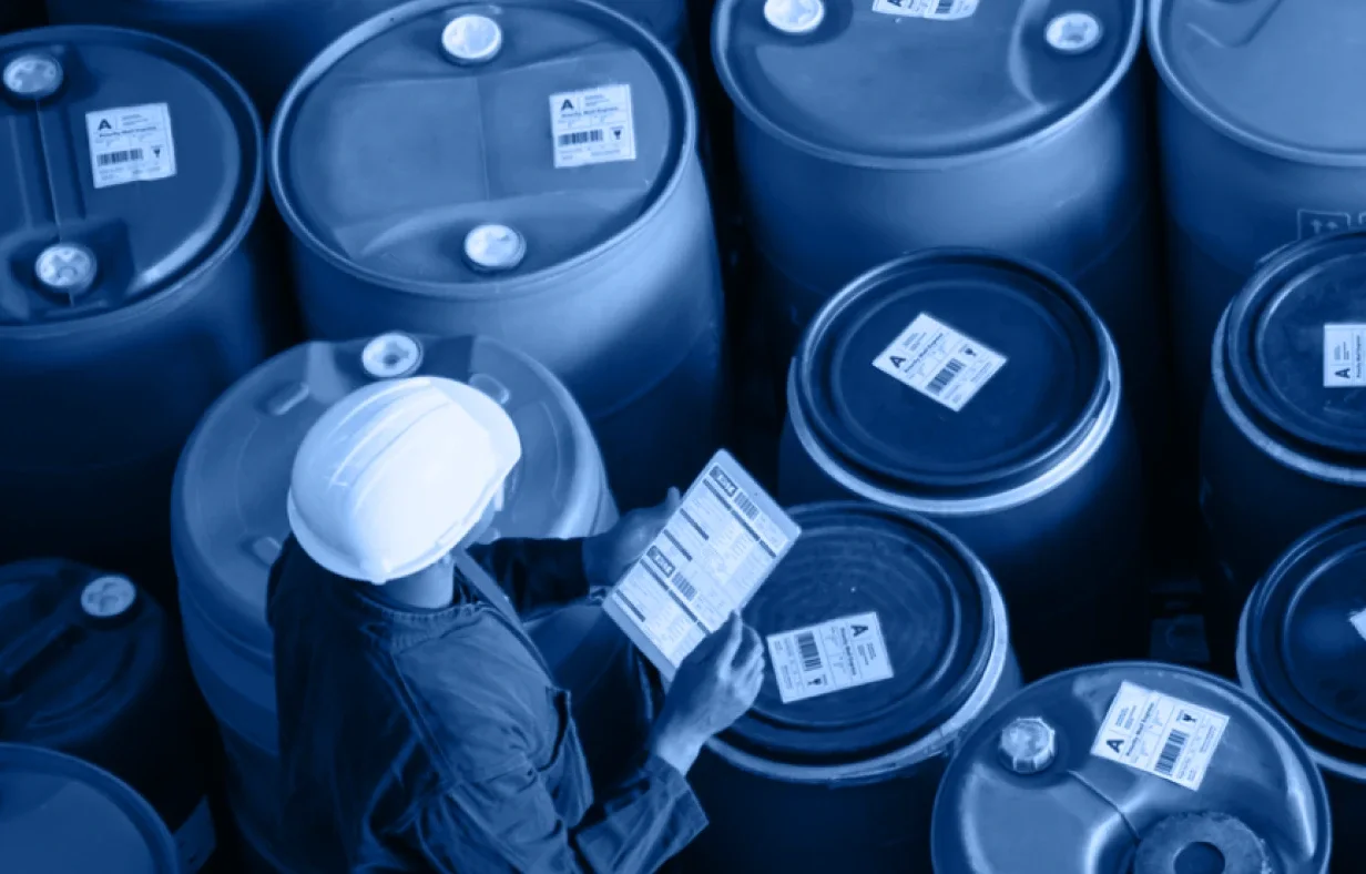 A person wearing a hard hat and work clothes inspects labels on large industrial barrels arranged in rows, ensuring safety or inventory details in an Industry Accordion storage facility.