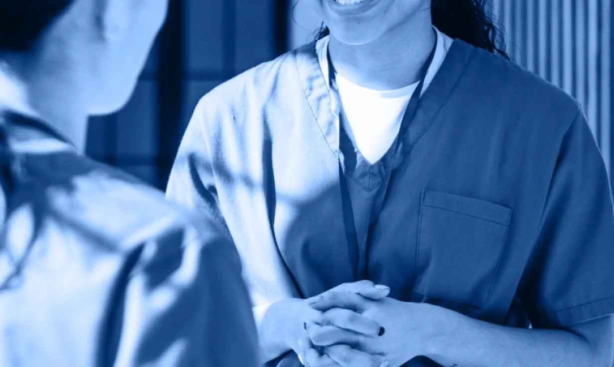 Two people in medical scrubs are standing and talking, with one smiling and clasping their hands together. The blue-tinted image captures a warm, home-like camaraderie often seen in the healthcare industry accordion of daily routines.