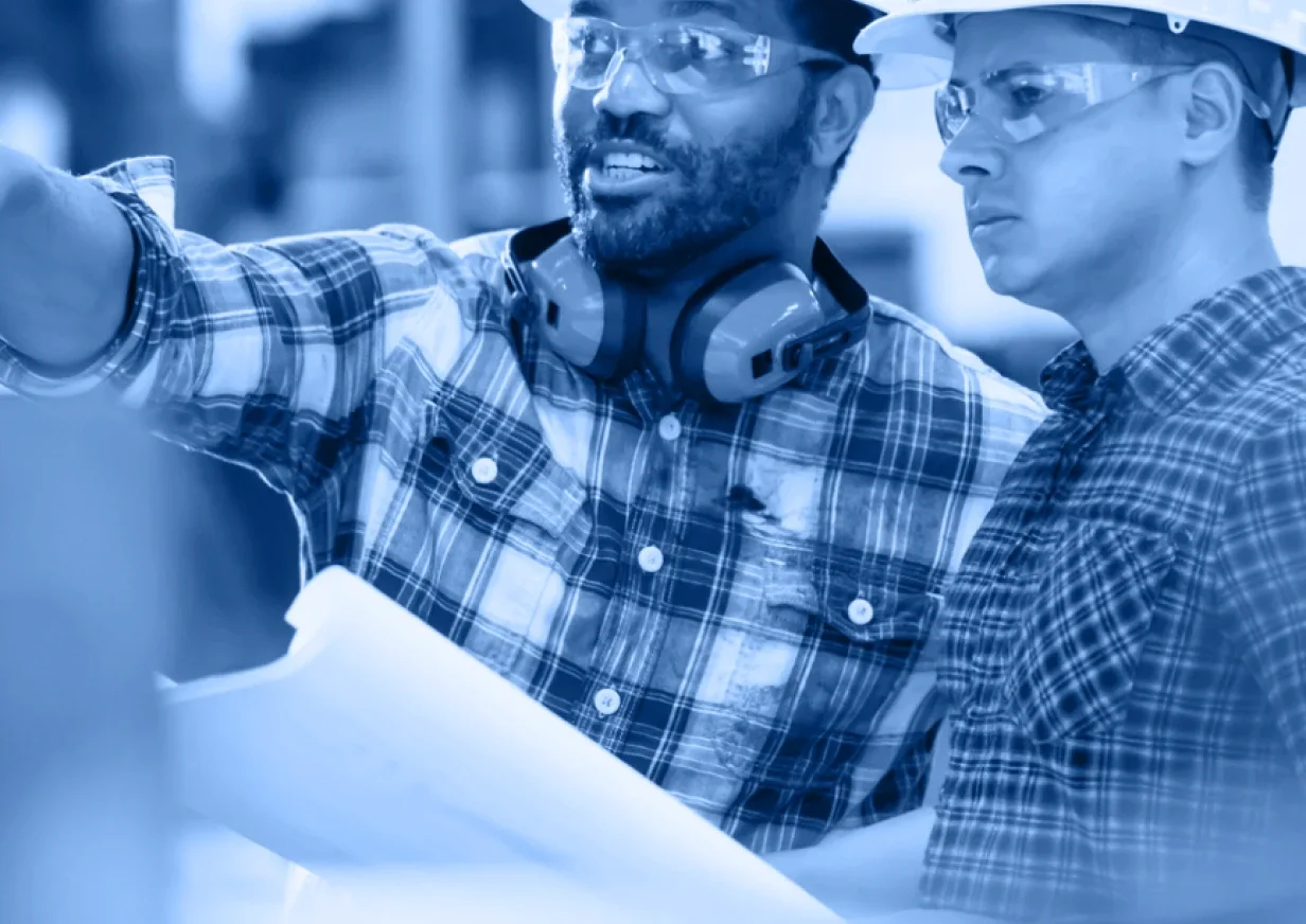 Two people wearing plaid shirts, safety glasses, and hard hats look at blueprints on a worksite. One points forward while the other listens, both engaged in discussion. The image has a blue tint, highlighting teamwork in an Industry Accordion Home project.