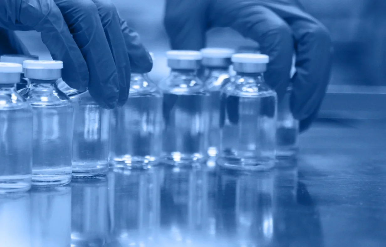 A gloved hand arranges several clear glass vials filled with liquid on a reflective surface in a laboratory setting, evoking an industry accordion of precision and order, with a blue color tone throughout the image.