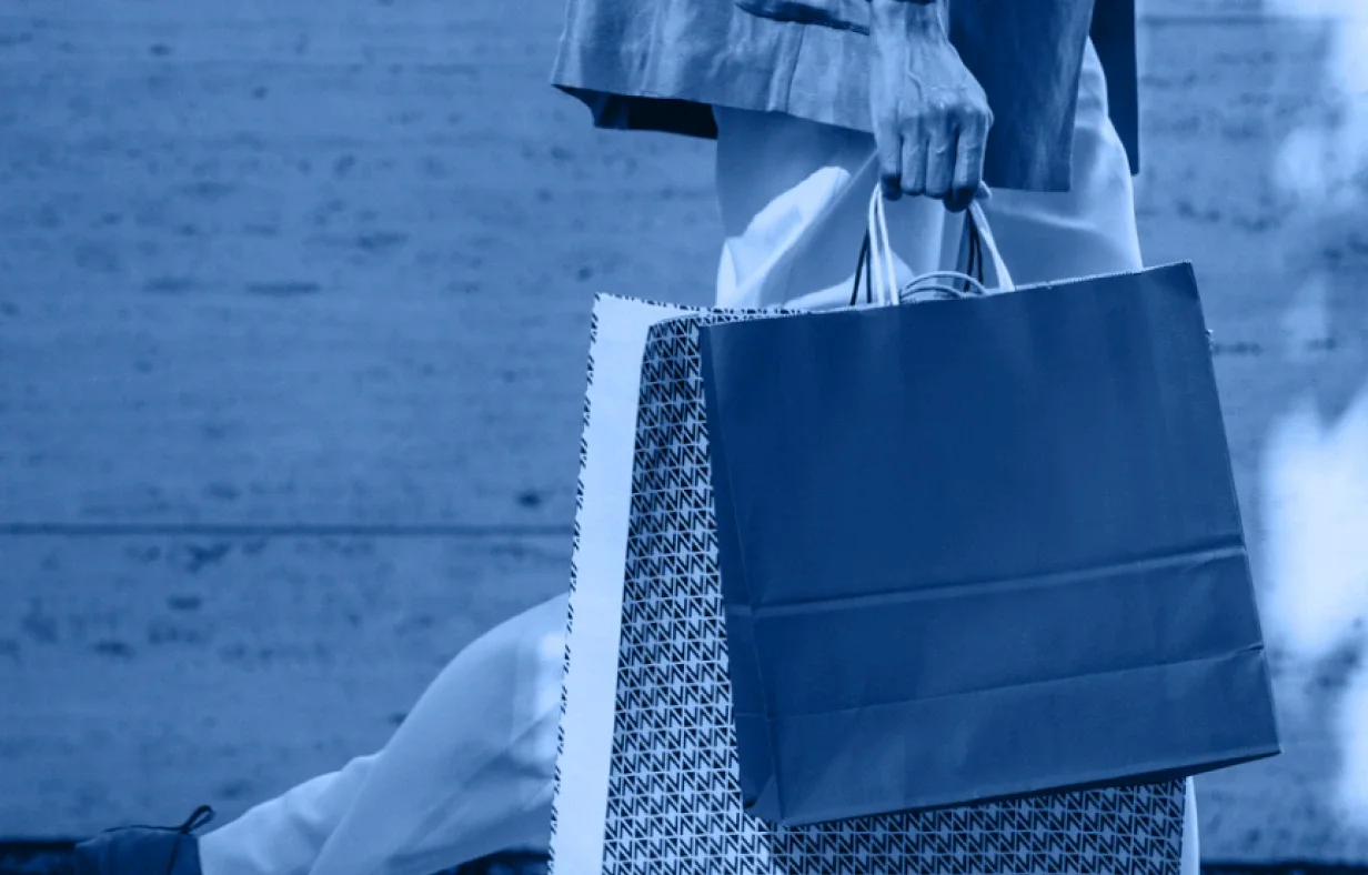 A person walks while holding several shopping bags in one hand, their lower body and hand captured against a plain background. The blue-tinted photo offers a modern take on Industry Accordion fashion trends.